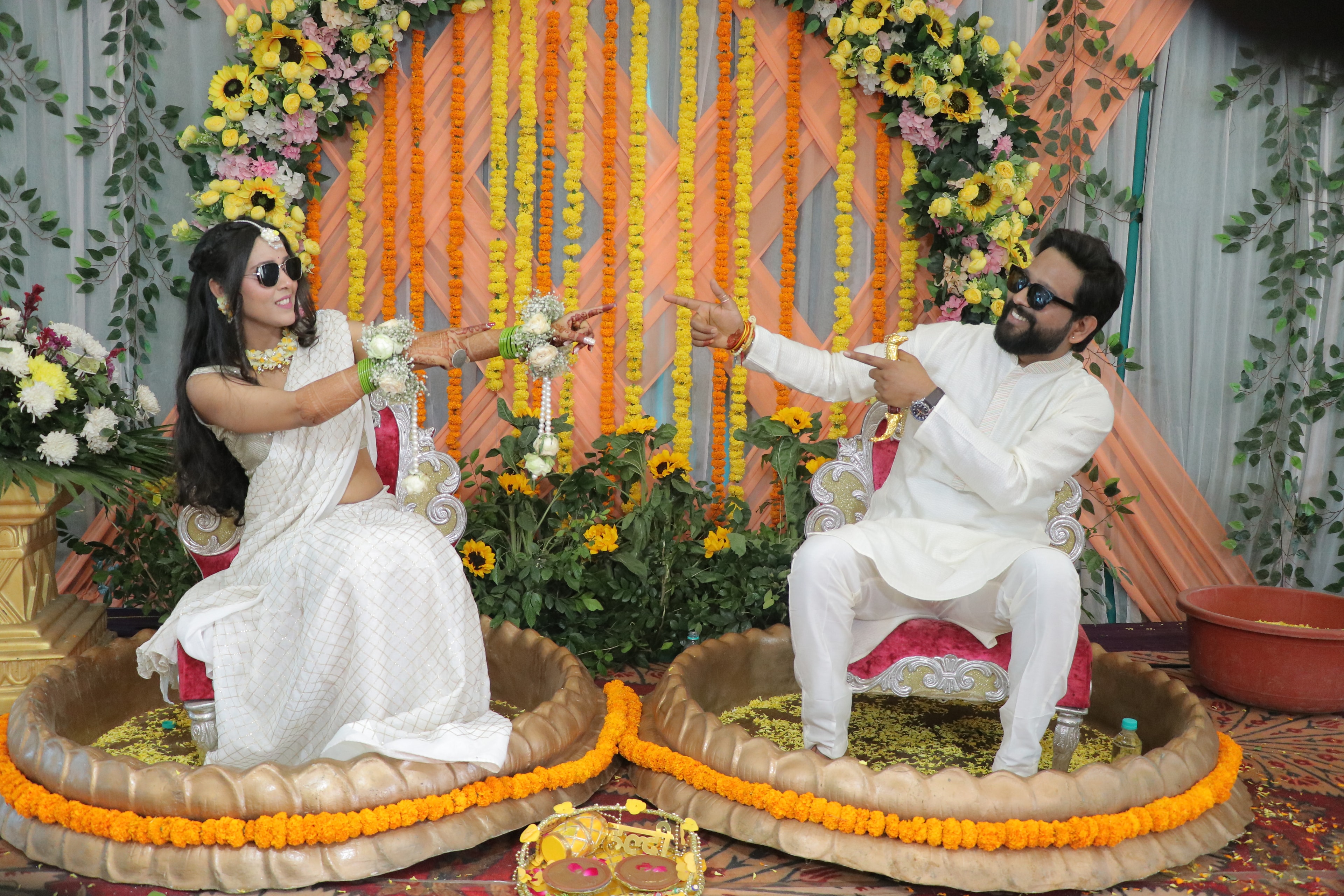 Bride and groom in white attire playfully pointing at each other on ornate thrones at a joyful Haldi ceremony in Agra, framed by marigold garlands, sunflowers and a vibrant floral backdrop — 250 pax destination wedding